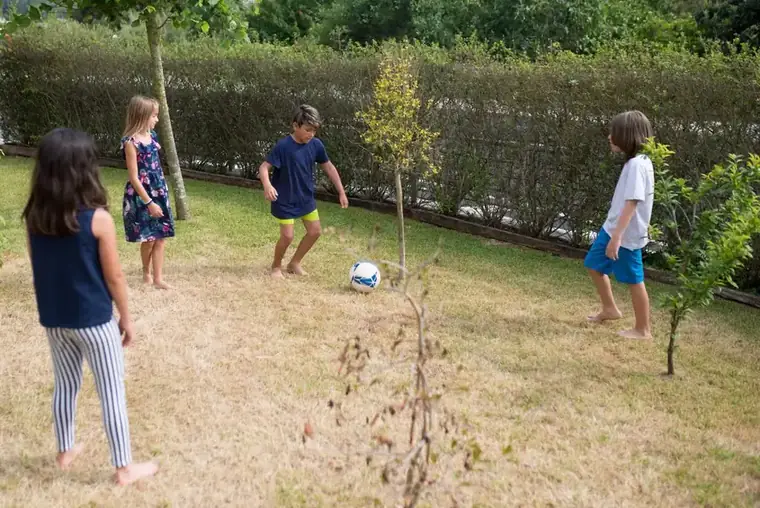 Four children playing football on a dry, worn summer lawn in front of a tall hedge