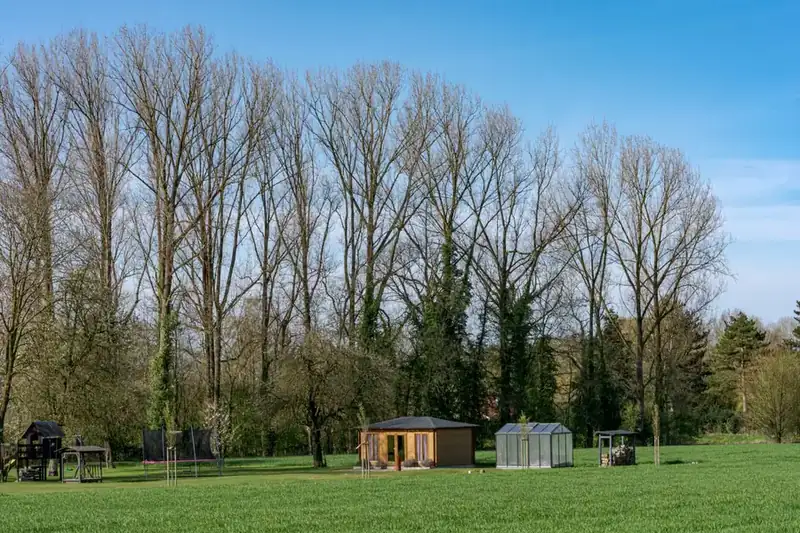 Timber garden cabin and a glass greenhouse at the far end of a large lawn, backed by tall bare trees