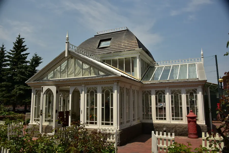 White-framed greenhouse standing in a garden with mature trees behind