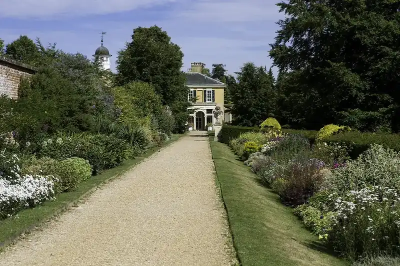 Long gravel path between deep herbaceous borders and lawn, leading to a large country house