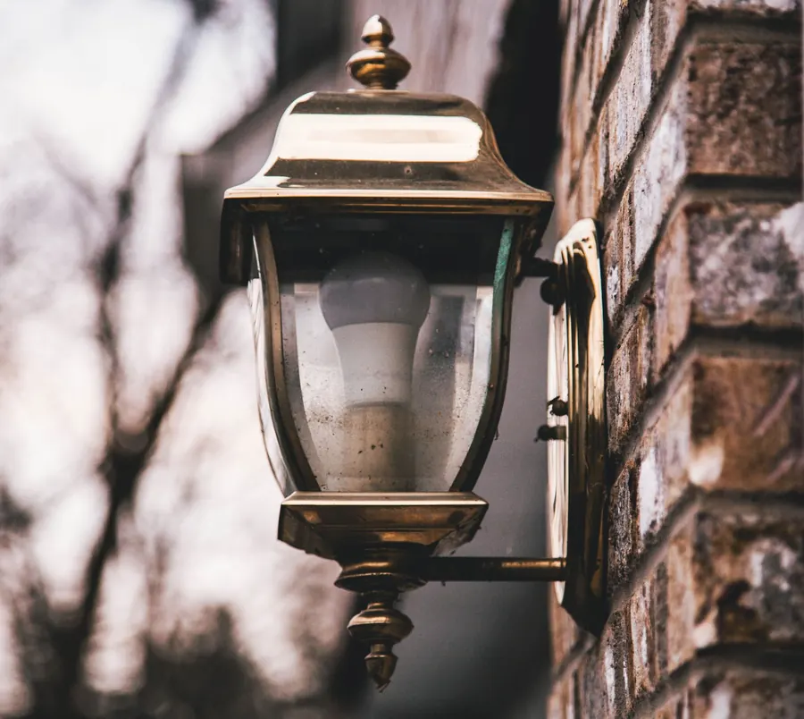 Brass mains-voltage wall lantern fixed to a brick house wall, the type of outdoor fitting that falls under BS 7671 wiring rules in a Hertfordshire garden