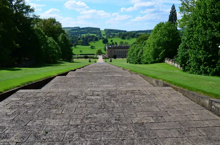 Broad natural stone path running straight between lawns towards a country house, the kind of setting-out a garden designer specifies down to joint lines and falls