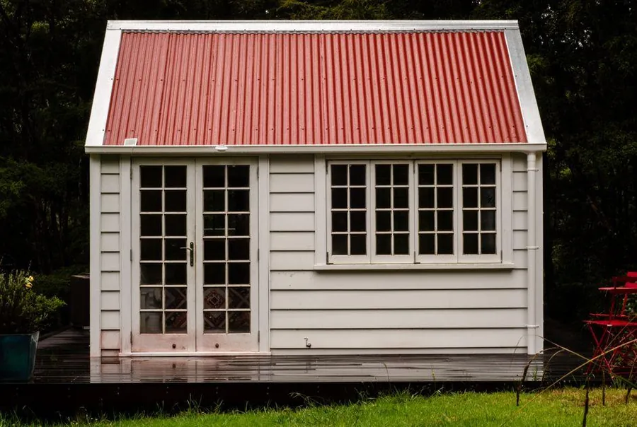 Small white garden room with a pitched red roof and glazed doors standing on low decking beside a lawn
