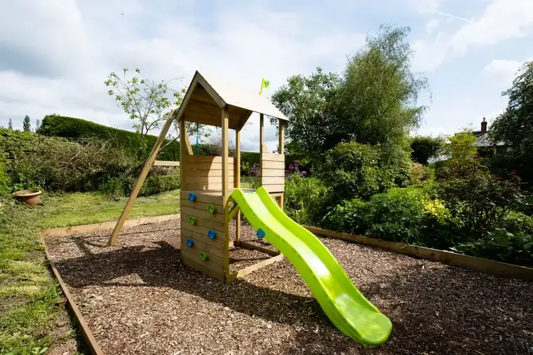 Wooden play tower with a green slide standing on a bed of bark chip edged with timber