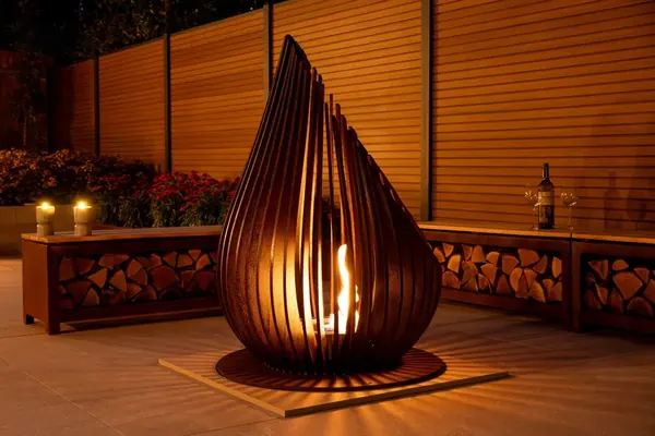 Sculptural steel fire pit glowing at night beside a slatted screen and log store